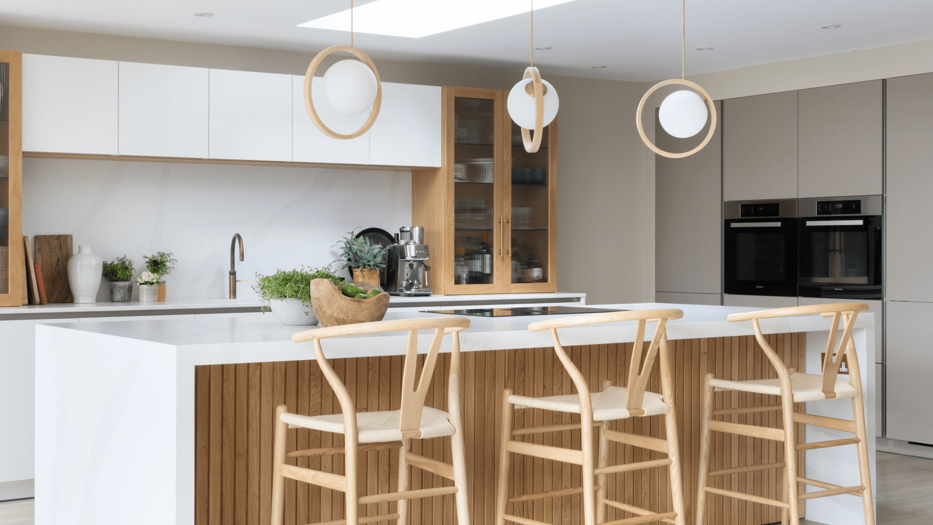 Modern kitchen with a perfect blend of style and function: A bright, airy kitchen featuring white cabinets with gold hardware, a marble waterfall island with bar stools, pendant lighting, open shelving displaying styled dishware, fresh herbs in windowsill planters, and a decorative tile backsplash. Natural light streams through large windows, highlighting both beautiful design elements and practical storage solutions.